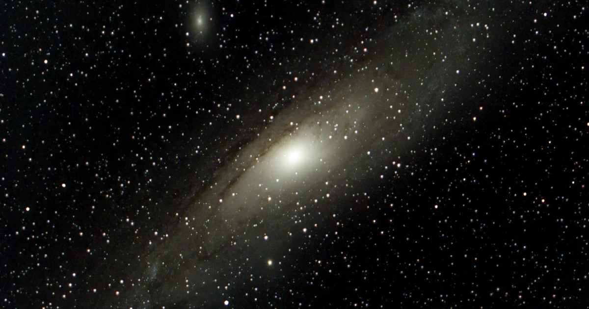 image of the andromeda galaxy: A disk of light is floating between small and big stars. The disk is bright in the center, has rings and fades to darkness at the edges.